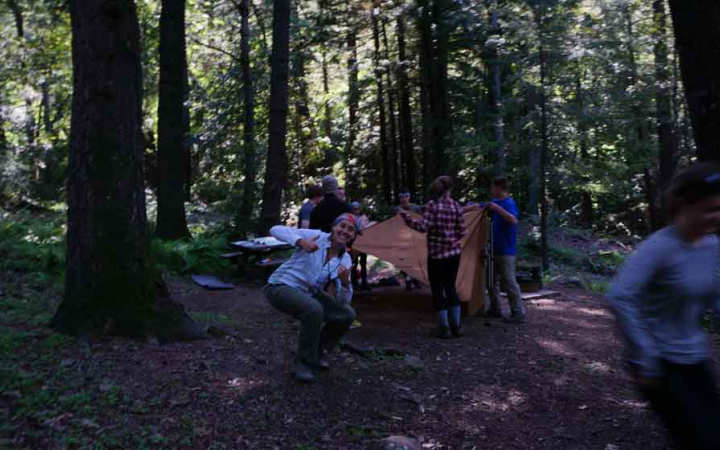 A group of students work to set up camp in a wooded area. A person in the foreground is giving the camera a thumbs up.